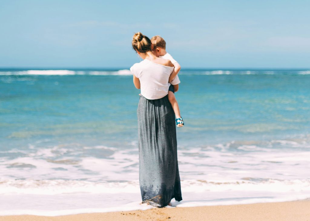 Mutter trägt ihr Kind am Strand und blickt aufs Meer, Sinnbild für verbundene Familienberatung in Buchholz in der Nordheide